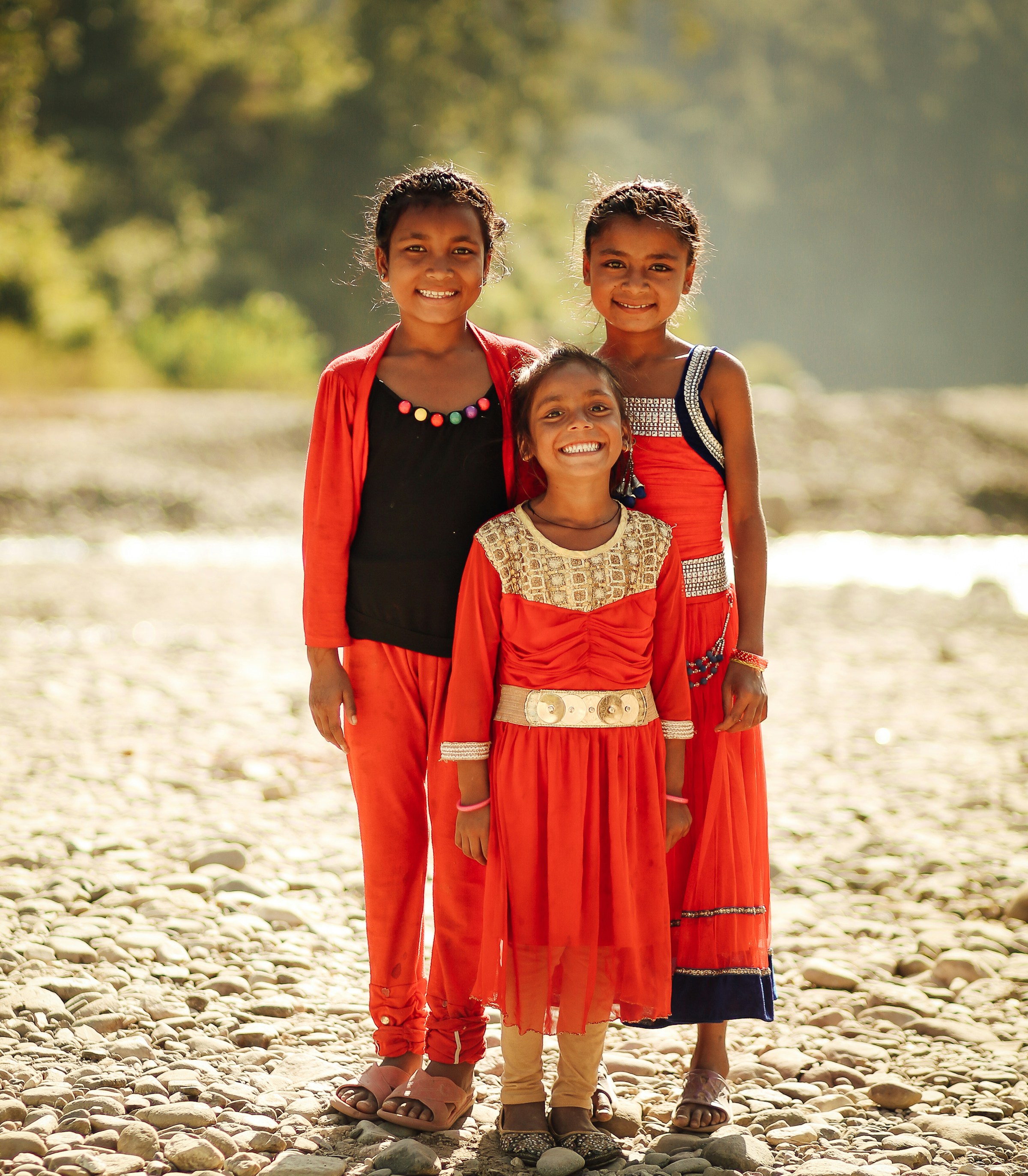 Three children standing together outdoors in warm sunlight, smiling.
